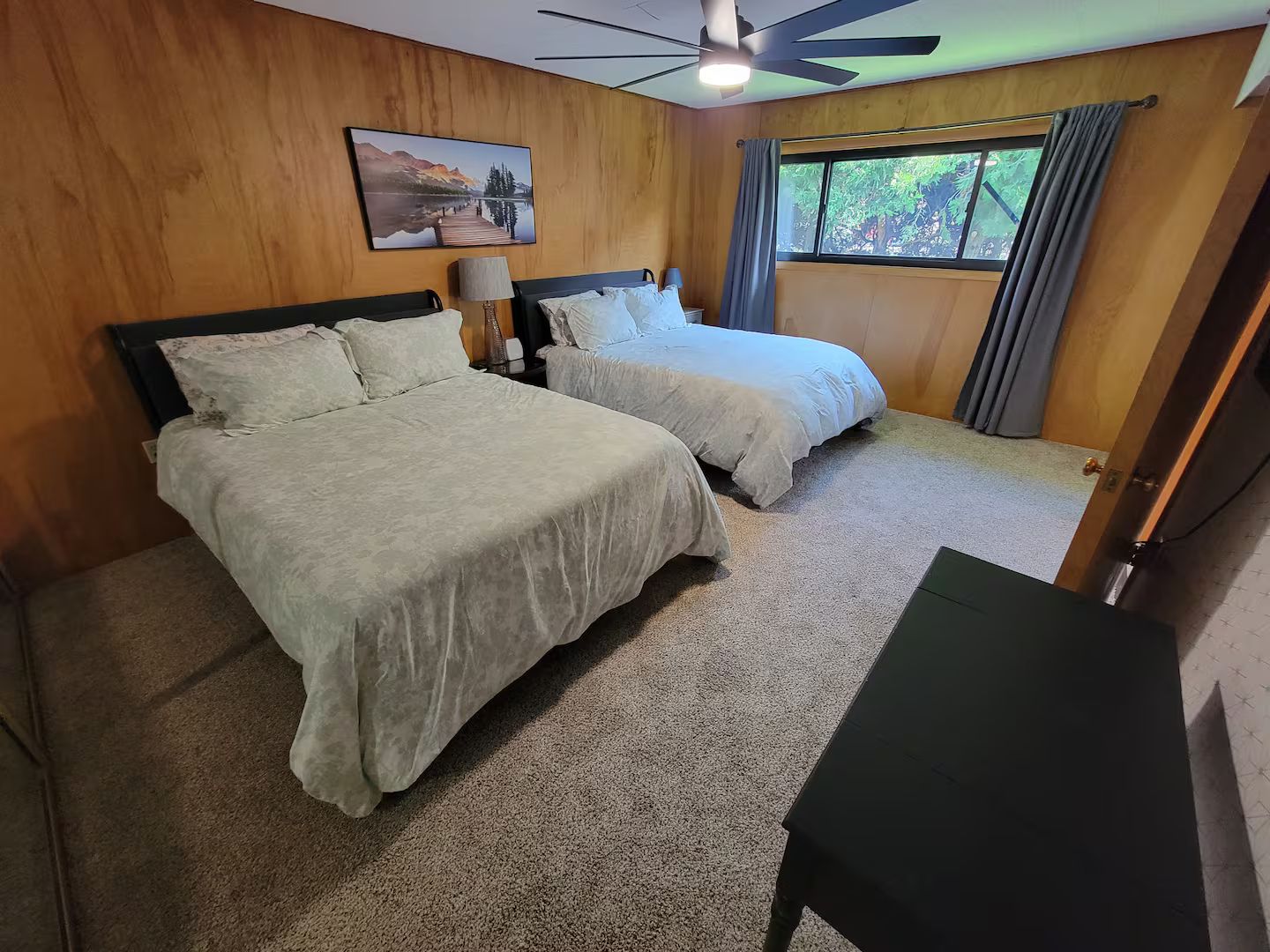 A hotel room with two beds, a ceiling fan, a window with curtains, and a painting on the wall.