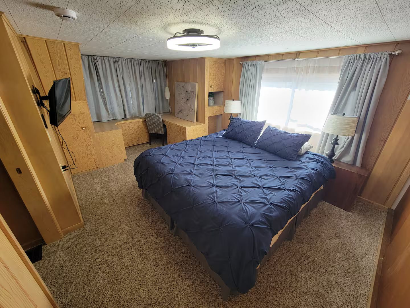A bedroom with a large bed, a desk, a chair, a television, and a window with curtains.
