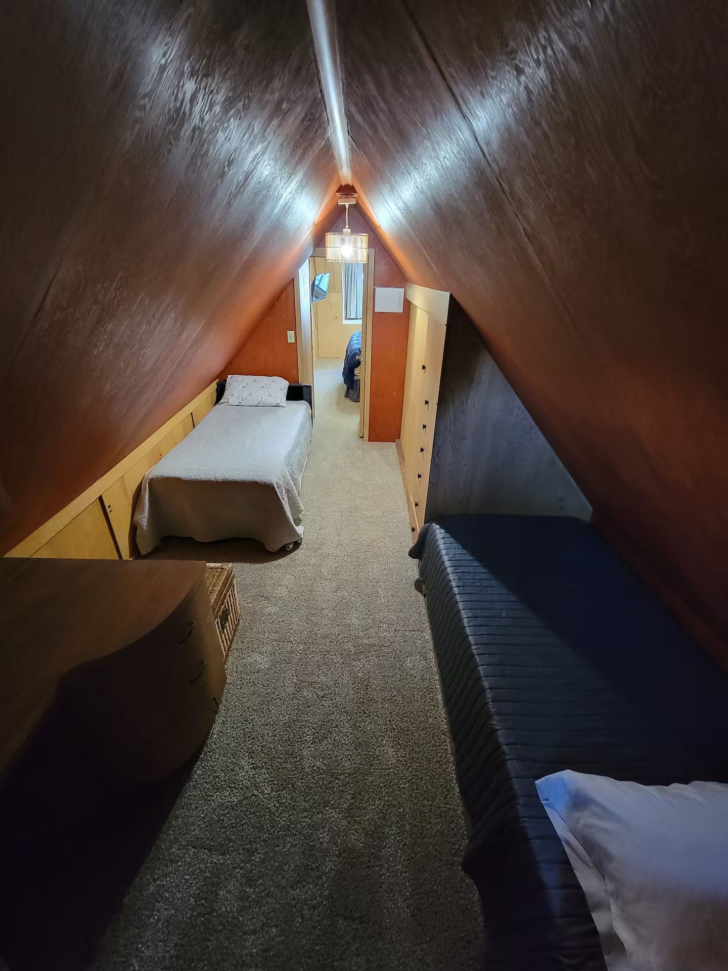 A narrow attic bedroom with two beds, a carpeted floor, and a hanging light fixture.