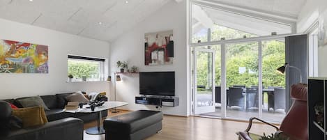 A modern living room with a flat-screen TV, a sofa, a coffee table, and a large window offering a view of the outdoors.
