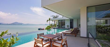 A modern outdoor pool area with a view of the sea, a dining table with chairs, and lounge chairs.