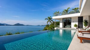 Ein moderner Infinity-Pool mit freiem Blick auf das Meer und die Berge, ein Balkon mit Glasbrüstung sowie eine Lounge-Bereich mit Liegestühlen.