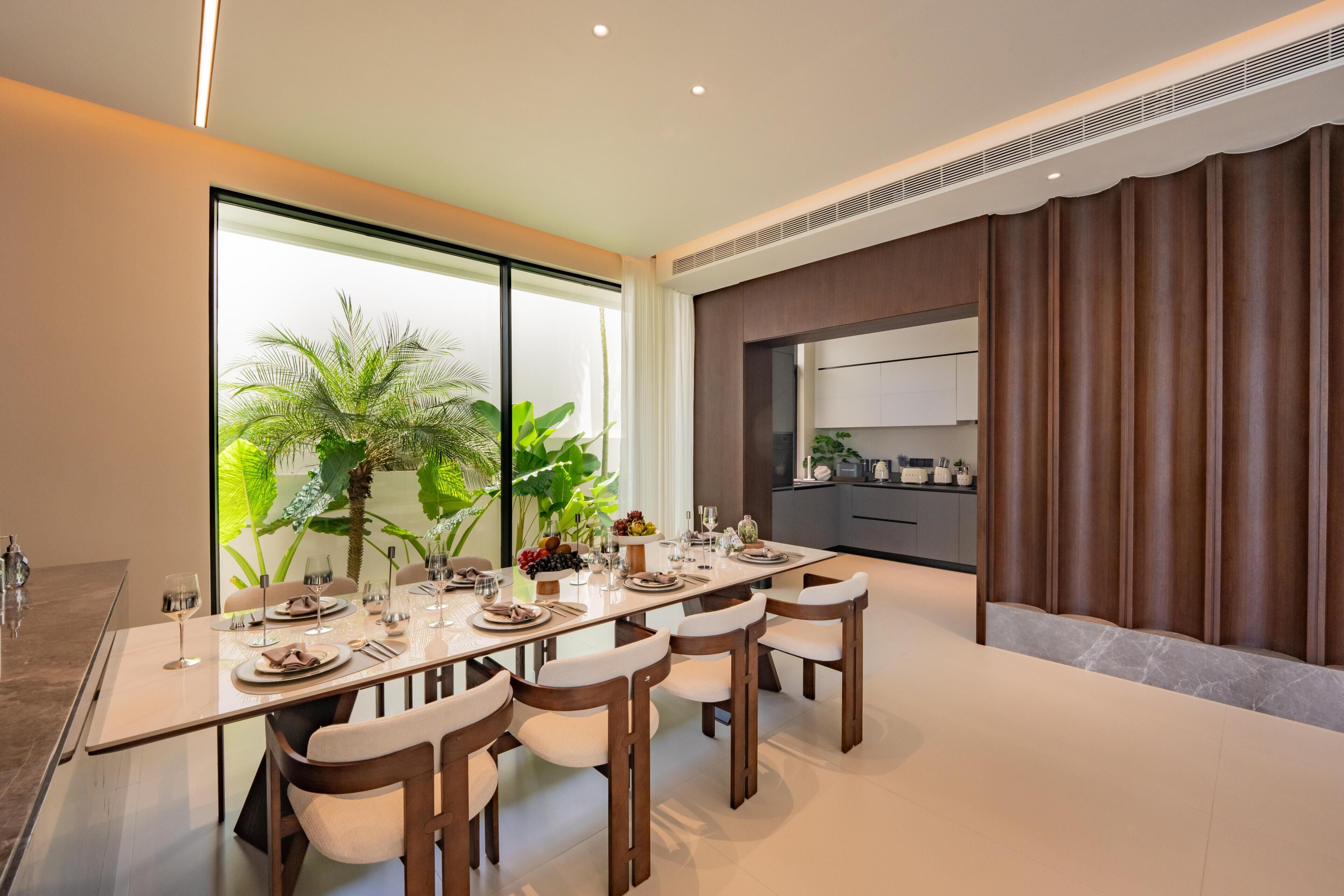 Uma sala de jantar moderna com uma mesa comprida, cadeiras e vista para uma cozinha e vegetação.