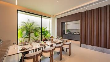 Uma sala de jantar moderna com uma mesa comprida, cadeiras e vista para uma cozinha e vegetação.