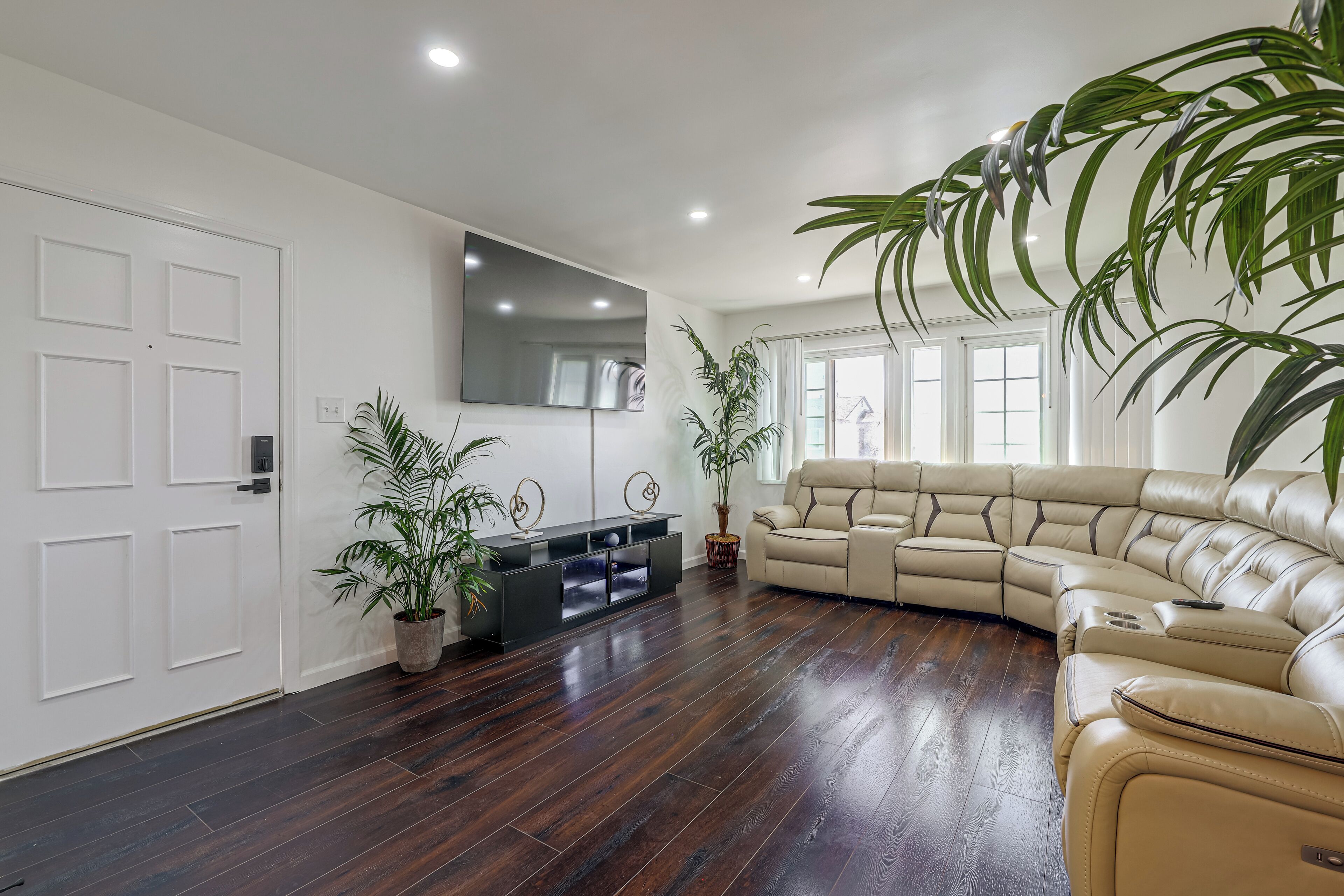A spacious living room with a large beige sectional sofa, dark wooden flooring, white walls, and potted plants.
