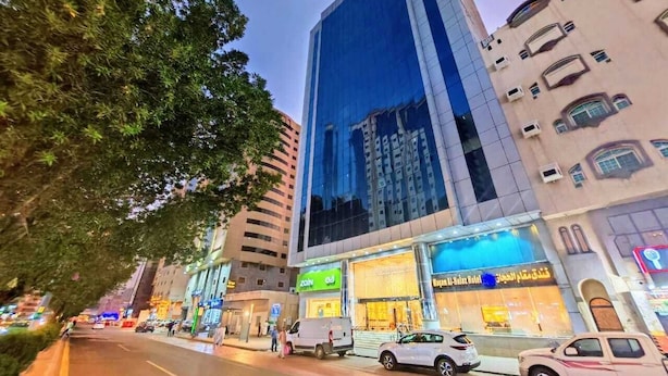 Hotel moderno com fachada de vidro, situado numa movimentada rua urbana com carros estacionados em frente.
