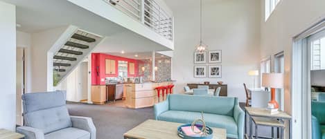 Un espacio habitable amplio con techo alto, una escalera que conduce a un nivel superior, una cocina con detalles rojos y un comedor con sofá azul.