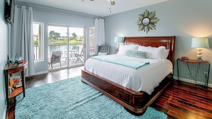 Une chambre avec un grand lit, un ventilateur de plafond, une vue sur l’extérieur et une petite table avec une lampe.