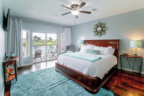 Een slaapkamer met een groot bed, een plafondventilator, uitzicht en een kleine tafel met een lamp.