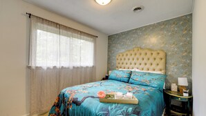 A bedroom with a floral bedspread, a tufted headboard, a bedside table with a lamp, and a window with sheer curtains. - Easy Raleigh Access! Quaint Apt w/ Patio (Garner)