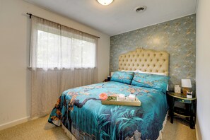 A bedroom with a floral bedspread, a tufted headboard, a bedside table with a lamp, and a window with sheer curtains. - Easy Raleigh Access! Quaint Apt w/ Patio (Garner)