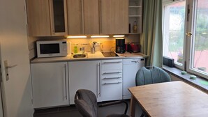 Une cuisine compacte équipée d’un four à micro-ondes, d’une machine à café et d’un évier. On y trouve une table à manger avec des chaises, ainsi qu’une fenêtre avec des rideaux.