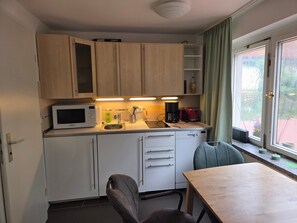 Eine kompakte Küche mit Mikrowelle, Kaffeemaschine und Spüle. Ein Esstisch mit Stühlen und ein Fenster mit Vorhängen sind ebenfalls vorhanden.