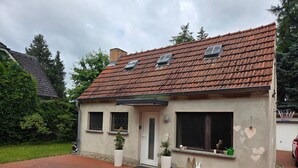 Une maison avec un toit recouvert de tuiles rouges, deux fenêtres avec volets, une porte blanche et un petit jardin agrémenté de plantes en pot.