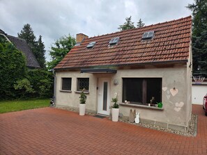 Ein Haus mit rotem Ziegeldach, zwei Fenster mit Fensterläden, eine weiße Tür und ein kleiner Garten mit Blumentöpfen.