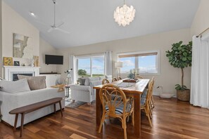 A spacious living room with a dining area, wooden flooring, and large windows offering a view.