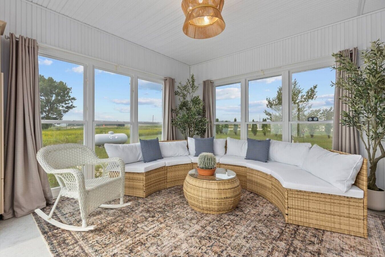 Ett rymligt rum med en böjd, vit soffa, ett flätat soffbord och en vit gungstol. Stora fönster ger utsikt över omgivningen.