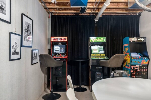 A room with arcade games, a table, and framed pictures on the wall.