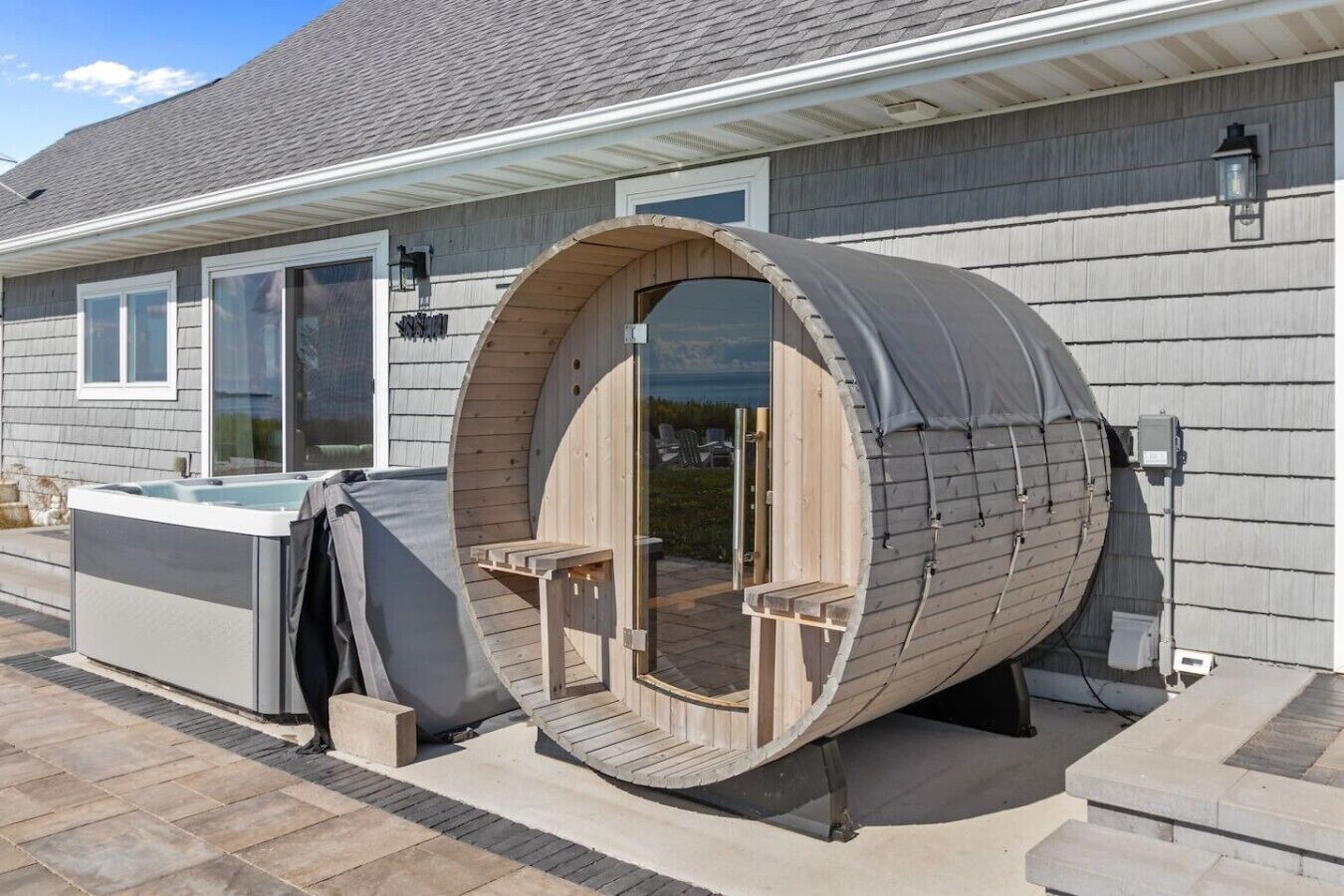 A sauna with a wooden exterior and a glass door, situated in a backyard with a hot tub and a house in the background.