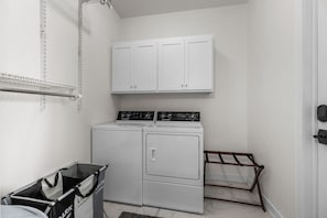 A laundry room with a front-loading washer and dryer, a drying rack, and a trash bin.