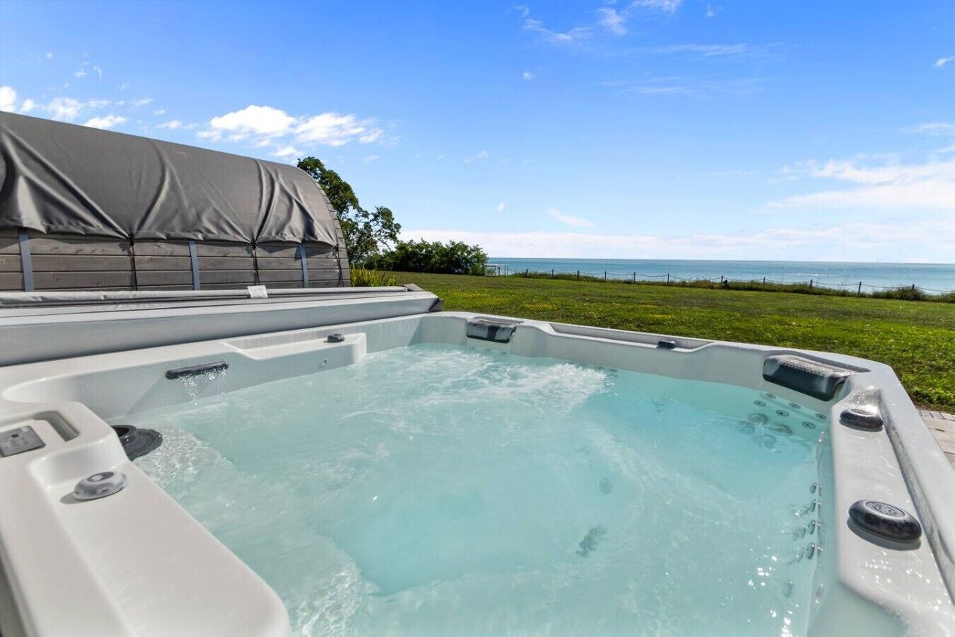 A hot tub with a view of the ocean, surrounded by a grassy area and a covered structure.