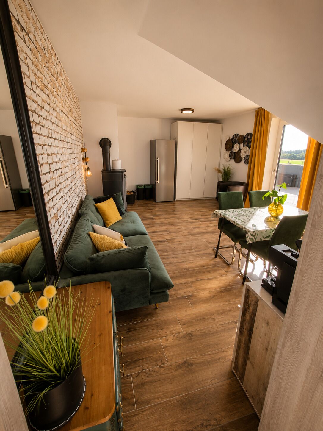 Une cuisine moderne avec un mur en briques, un coin repas avec une table et des chaises, et un canapé vert agrémenté de coussins jaunes.