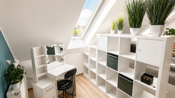 A modern, well-lit room with a sloped ceiling, a white desk, and a chair, accompanied by a large shelving unit and a potted plant.