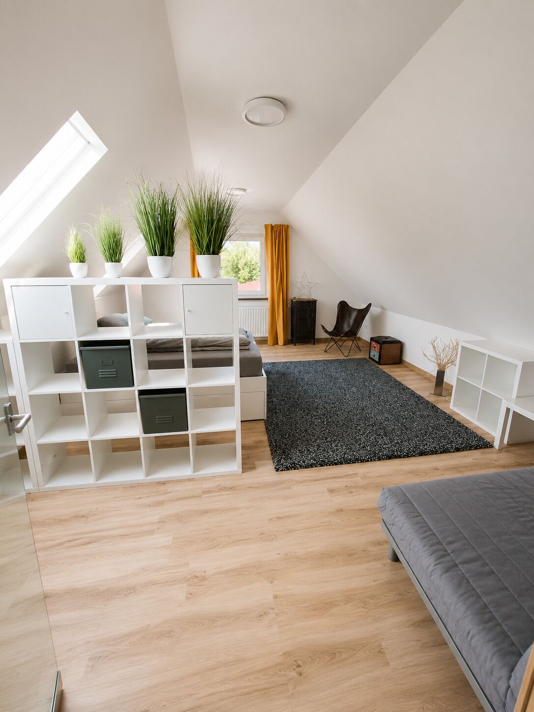 Ett modernt sovrum i loftstil med ett takfönster, inbyggd förvaring, en grå soffa och en svart matta.