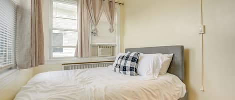 A bedroom with a bed, a window with curtains, a radiator, and a wall switch.