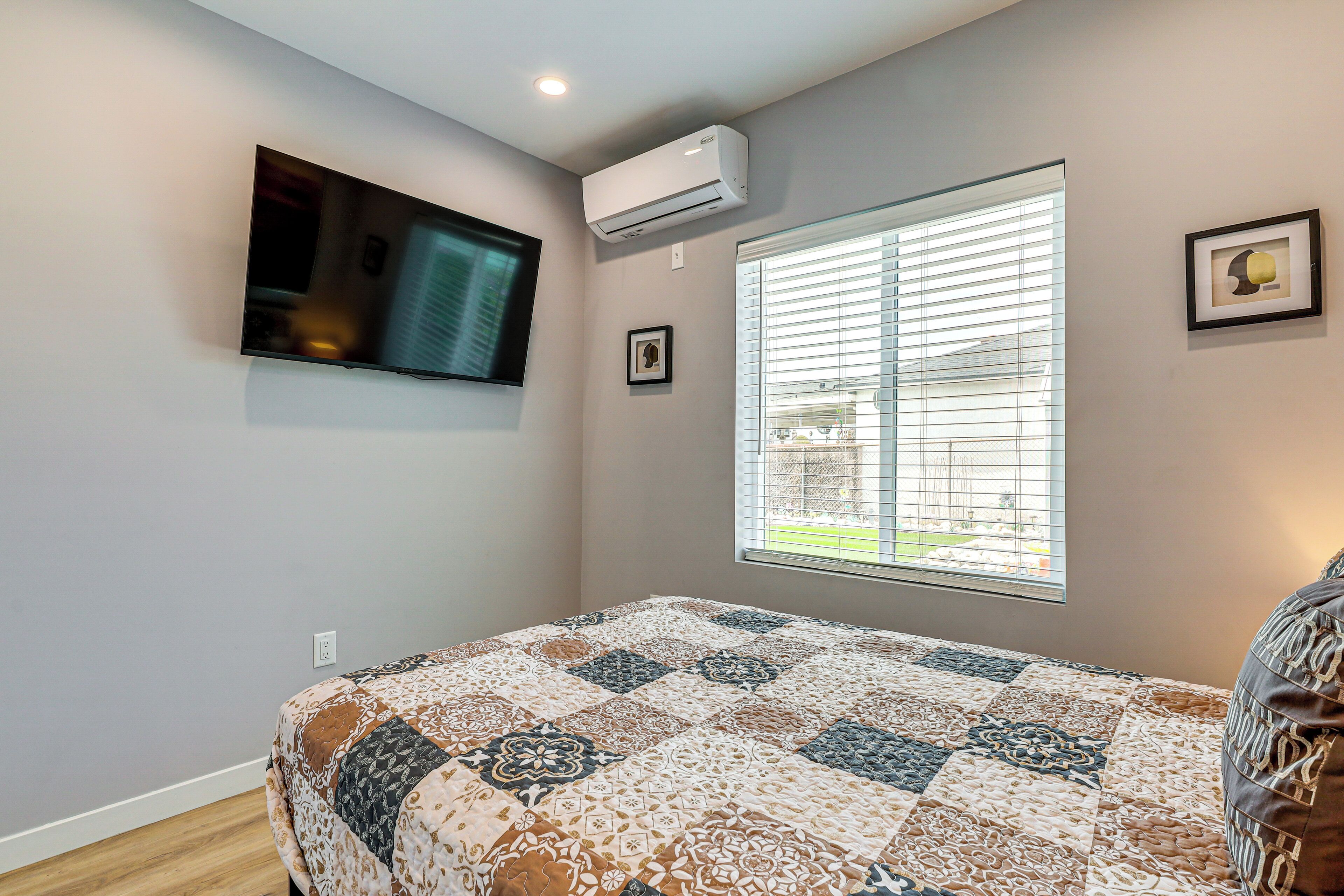 Un dormitorio con cama, televisor montado en la pared, aire acondicionado, ventana con persianas y un cuadro enmarcado.