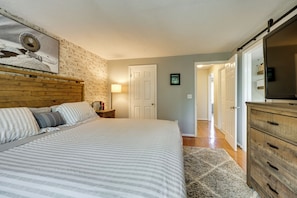 A bedroom with a wooden headboard, a bed with striped bedding, a nightstand with a lamp, and a dresser with drawers. - Home Gym & Office! Townhome Walk to Genesee River (Rochester)