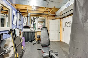 Een sportschool met een gewichtbank, diverse fitnessapparaten en een deur met de tekst 'GYM OPEN 24/7 NO EXCUSES!'