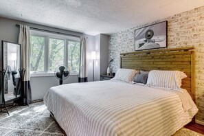 A bedroom with a large bed, a wooden headboard, a window with curtains, a standing fan, and a mirror. - Home Gym & Office! Townhome Walk to Genesee River (Rochester)