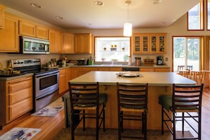 A kitchen with wooden cabinets, a central island with bar stools, a built-in oven, and a window with plants. - PNW Island Escape! Golf & Unwind in Nature (Everett)