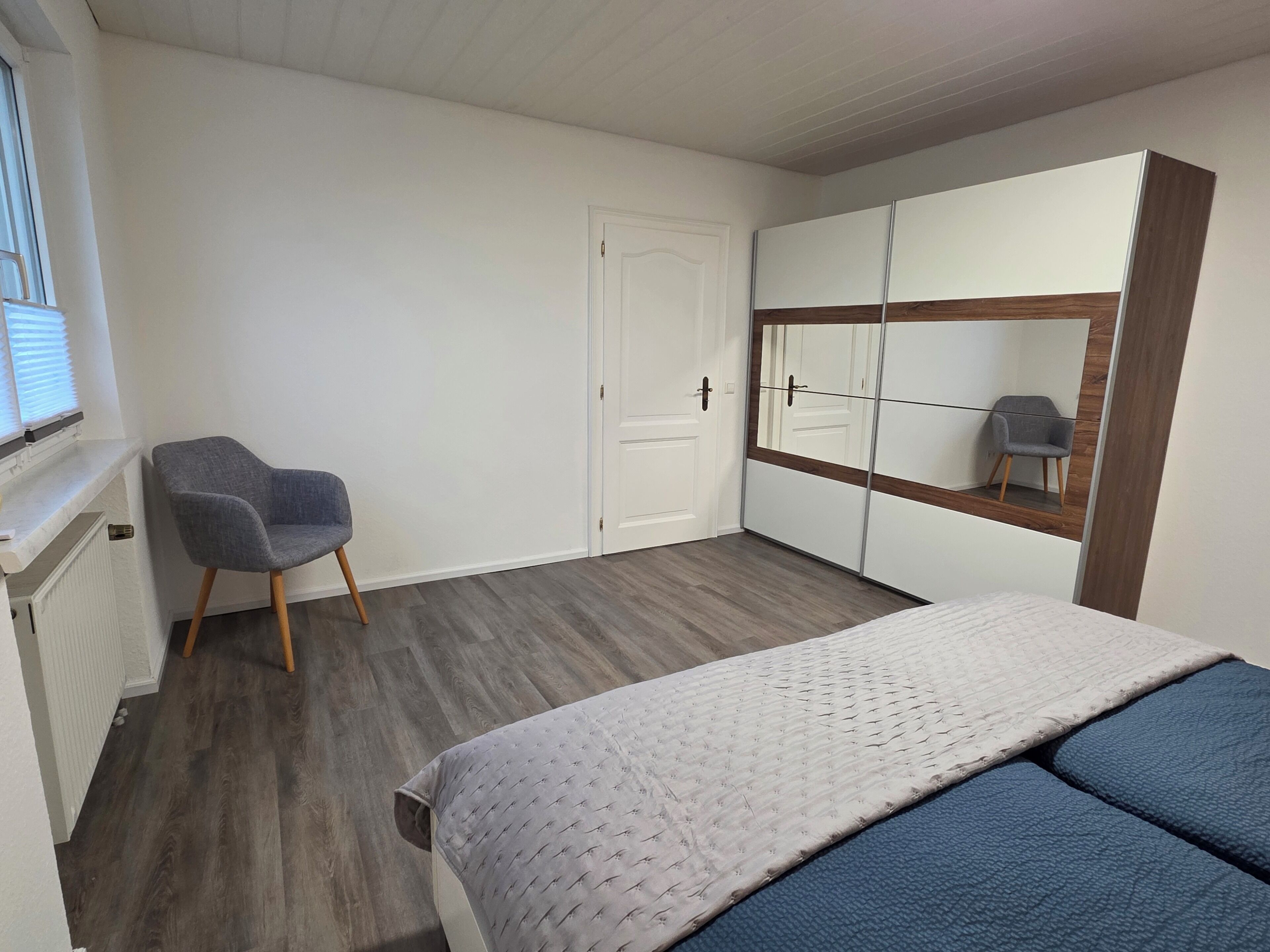 Un dormitorio con cama, una silla, armario y espejo.