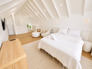 Ein modernes Schlafzimmer mit schrägverlaufender Decke, einem Holztisch, einem Bett mit weißer Bettwäsche und einem kleinen runden Hocker.