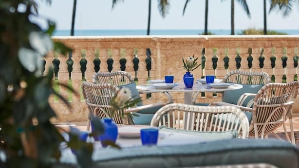 An outdoor dining area with wicker furniture and blue table settings, overlooking a beach with palm trees. - Four Seasons Hotel at The Surf Club.  (Surfside)