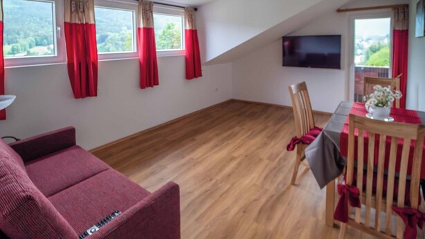 Una habitación con piso de madera, un sofá, una mesa de comedor con sillas y un televisor montado en la pared.