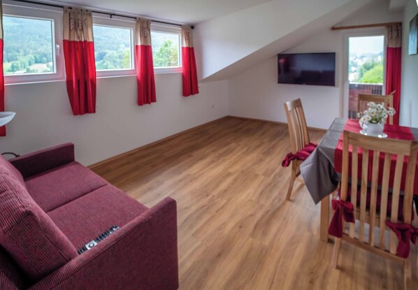 Een kamer met houten vloer, een bank, een eettafel met stoelen en een aan de muur bevestigde televisie.