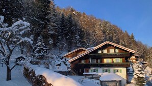 A wooden chalet with a sloped roof, surrounded by snow-covered trees and a stone wall. - Vrbo Property (Berchtesgaden)