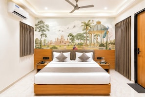 A hotel room with a bed, a nightstand, a wall-mounted air conditioner, a ceiling fan, and a mural depicting a historical building. - Hotel Teerth Dwarka (Dwarka)