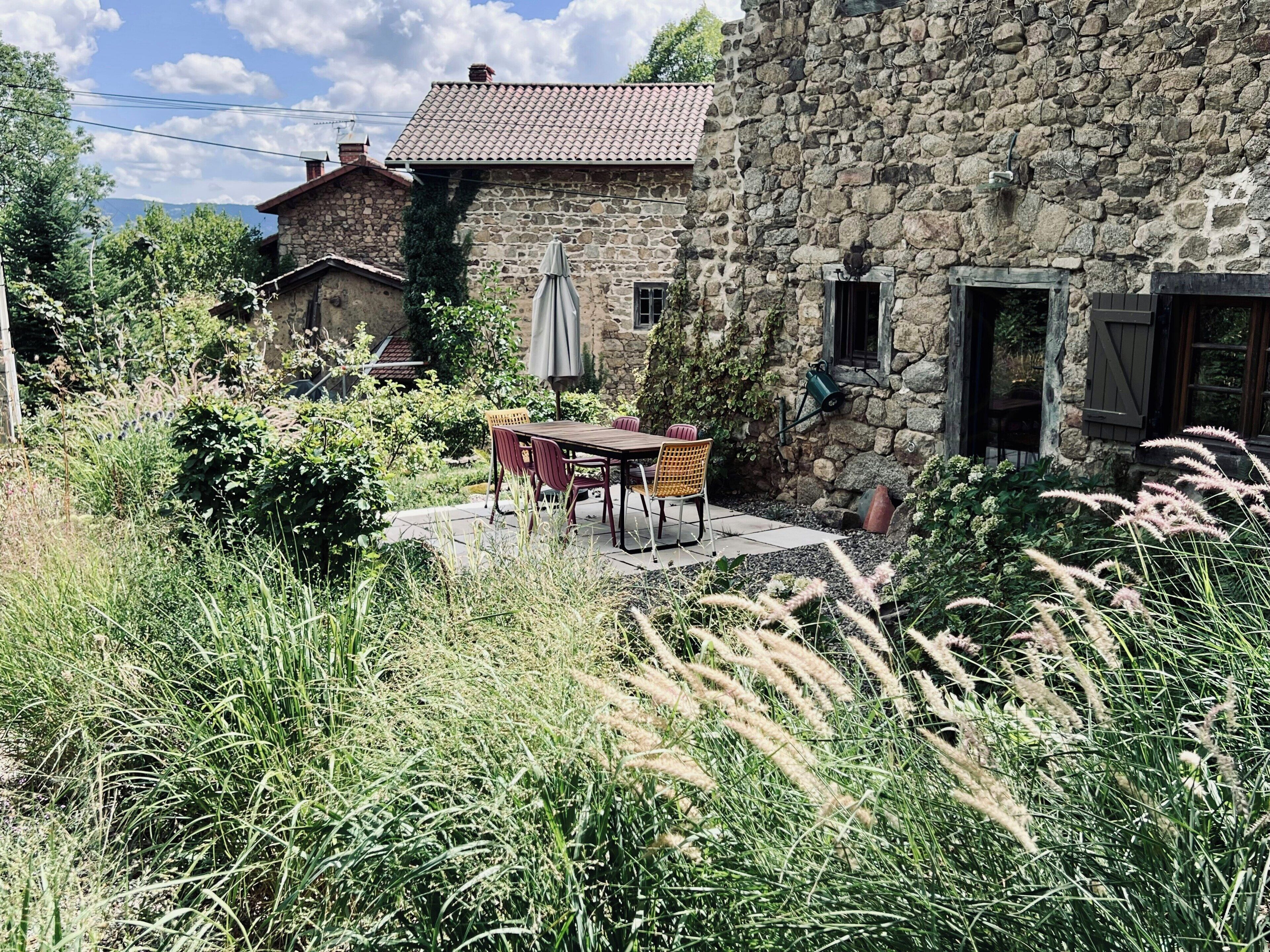 A stone building with a tiled roof, a patio with a table and chairs, and surrounding greenery.