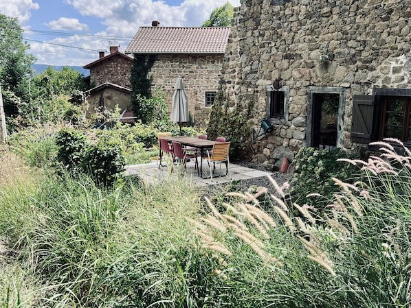 A stone building with a tiled roof, a patio with a table and chairs, and surrounding greenery. - Stone house in the hills of the Livradois (Marsac-en-Livradois)