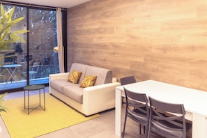 A modern living room with a sofa, a dining table, and chairs. There is a large window with a view of trees.