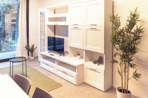 A modern living room with a flat-screen TV, white cabinetry, a dining area with a table and chairs, and a large potted plant.