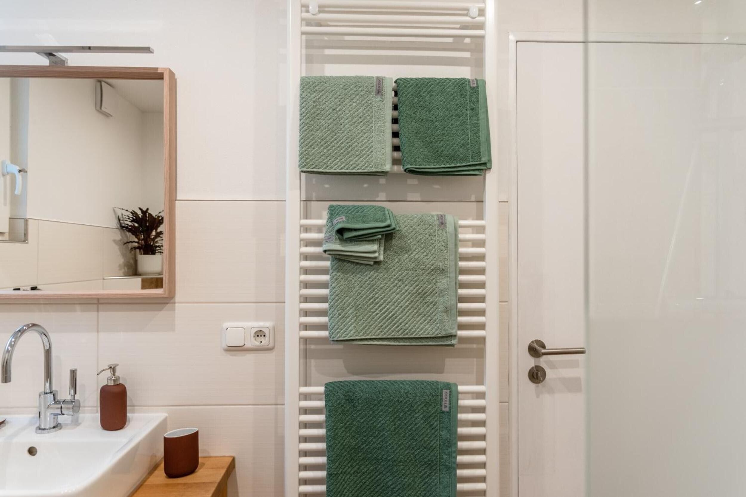 Ein Badezimmer mit einem weißen Waschbecken, einem Spiegel, einem Handtuchhalter mit grünen Handtüchern, einem weißen Heizkörper und einer Tür.