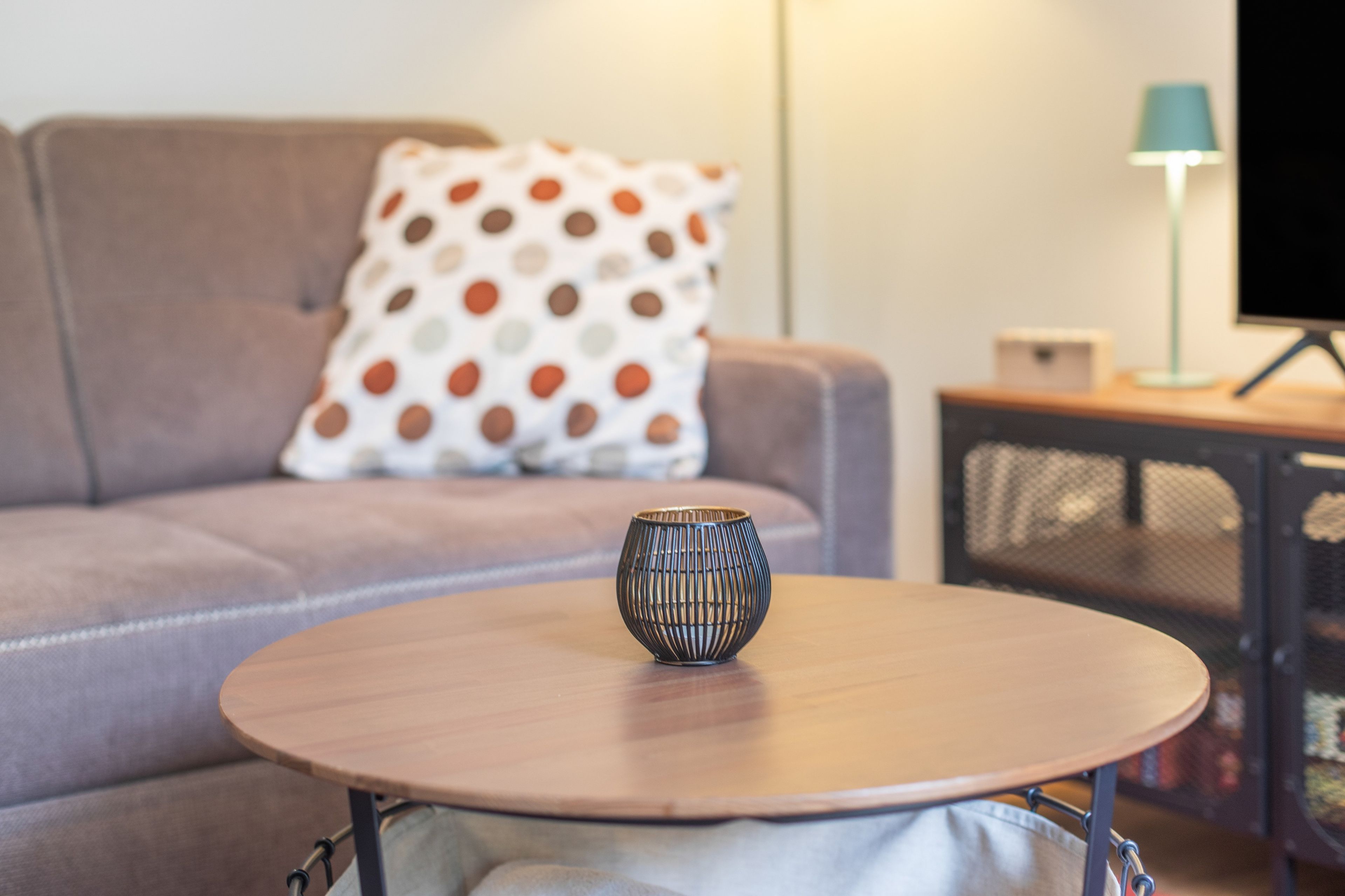 A round wooden table with a decorative vase in the center, a sofa with a polka dot pillow, and a lamp on a side table.
