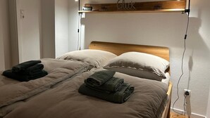 Une chambre avec deux lits, une tête de lit en bois, une étagère avec une horloge et une lampe fixée au mur.