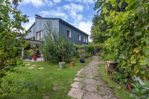 Un chemin de jardin mène à une maison à l’extérieur bleu, entourée de différentes plantes en pot et de verdure.