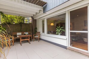 Un espace extérieur aménagé en terrasse, avec une table et des chaises en bois, un banc et une grande fenêtre donnant sur un jardin.